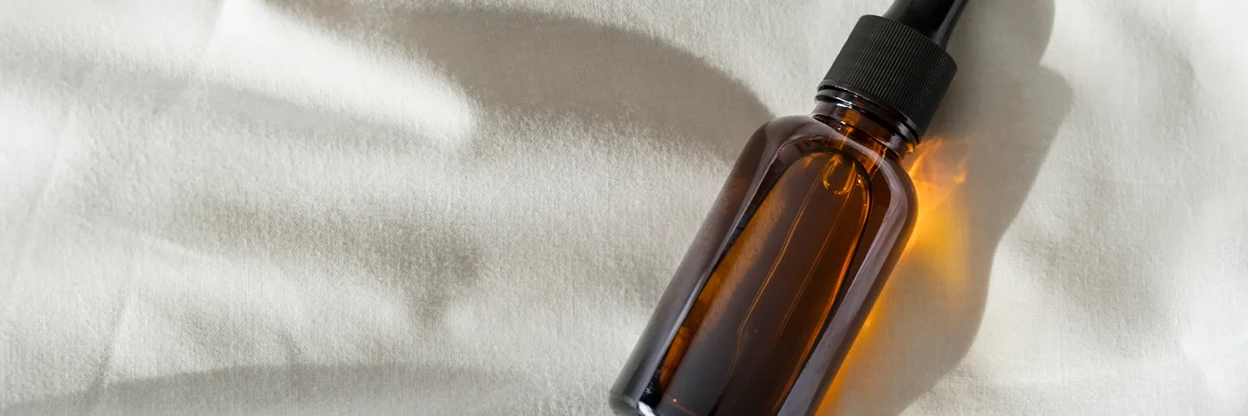 An unlabelled amber glass dropper bottle lying on softly folded white fabric in raking sunlight.