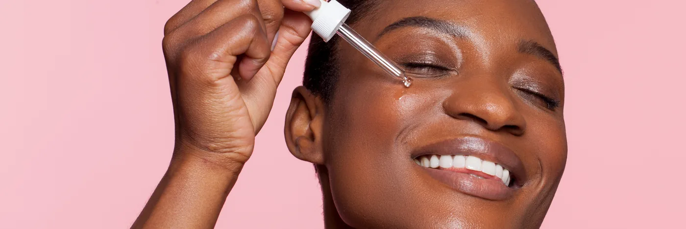 A smiling woman holding a glass pipette to her cheekbone, applying a drop of serum, against a pink background.