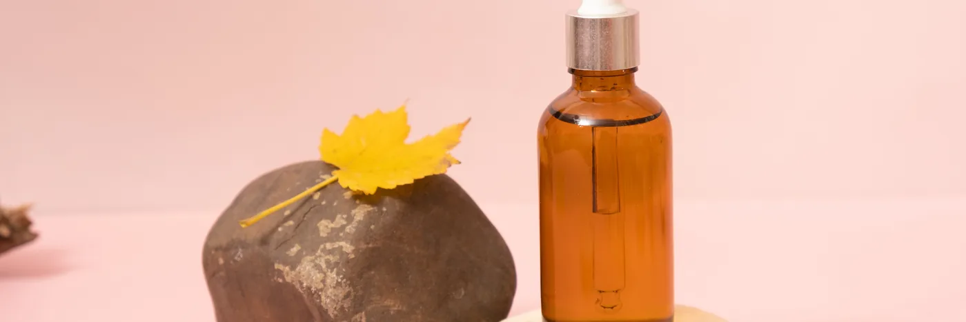 An unlabelled amber dropper bottle on a pale pink surface next to a smooth stone holding a single yellow leaf.