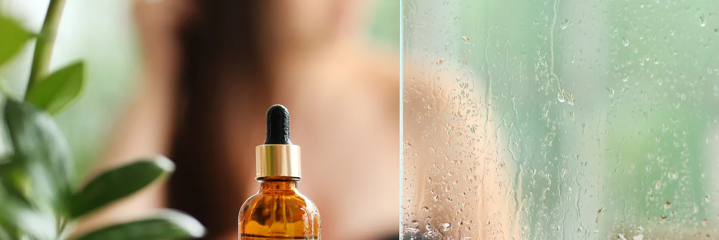 An unlabelled amber dropper bottle on a bathroom shelf beside a green plant, with fogged shower glass behind.
