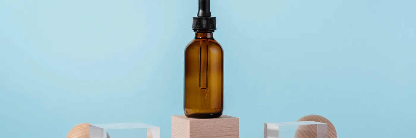 A single unlabelled amber dropper bottle standing on a pale wooden block against a soft blue background, with geometric props arranged around it.
