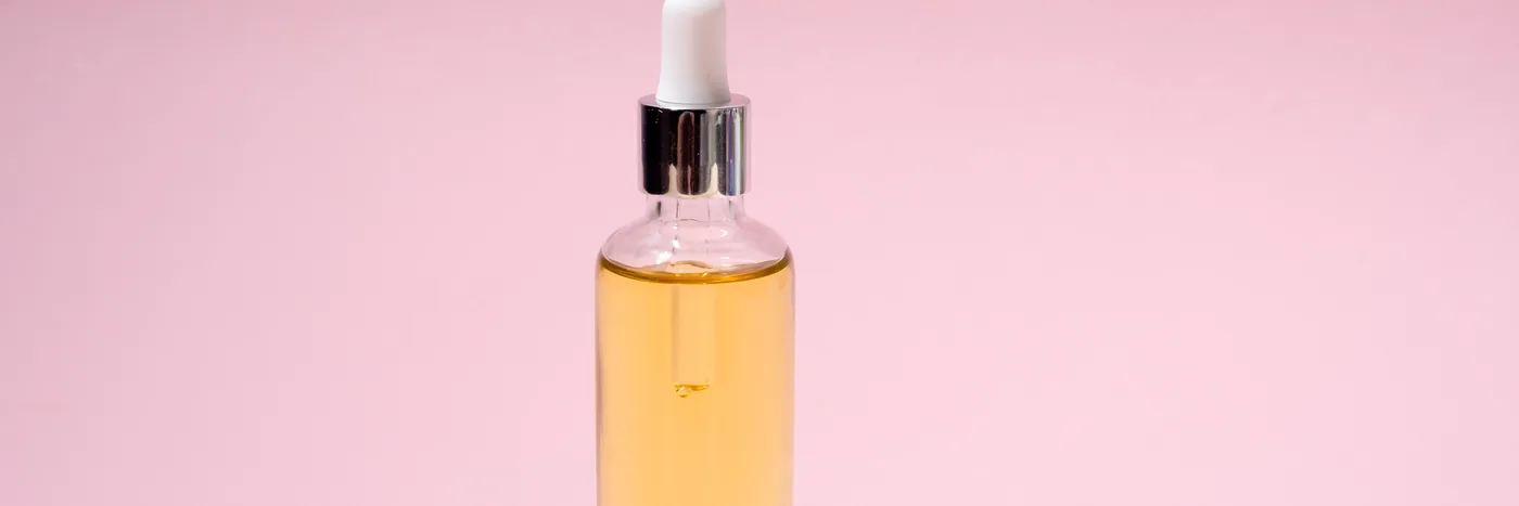 A tall unlabelled glass serum bottle filled with pale gold liquid, lit against a pink background.