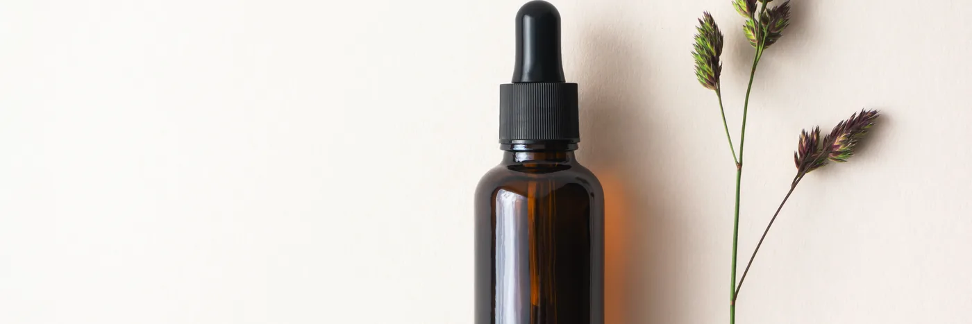 An unlabelled dark glass dropper bottle standing on a cream background beside a dried meadow grass stem.