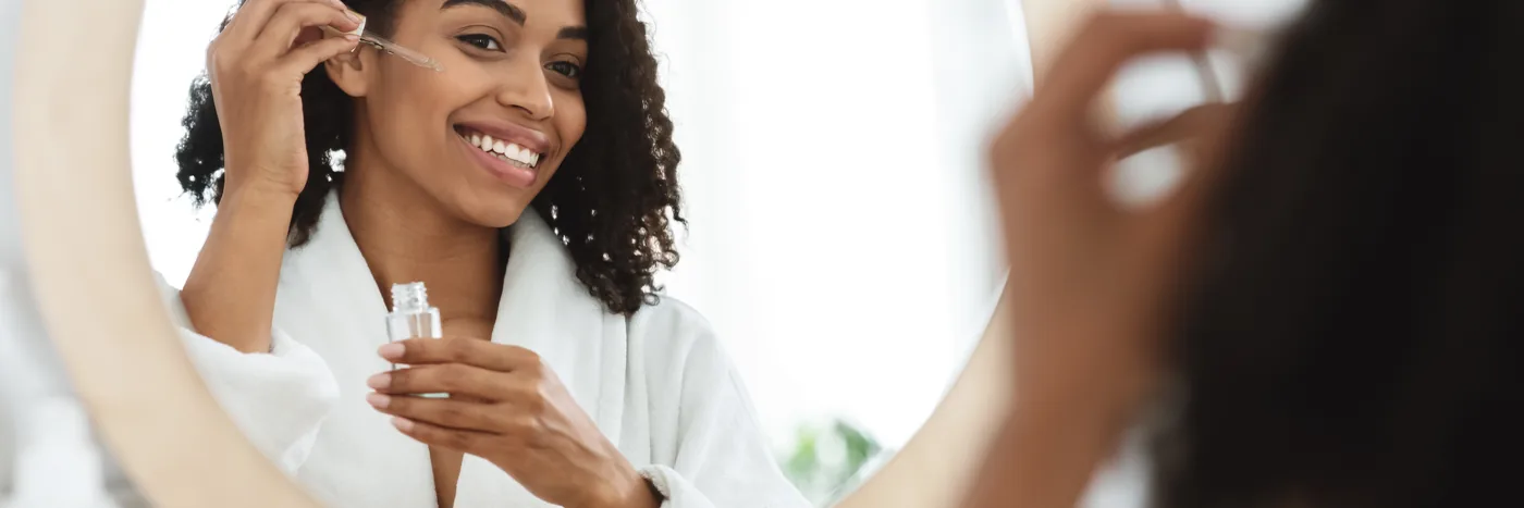 A woman in a white robe smiling into a round bathroom mirror while applying serum from a small bottle.