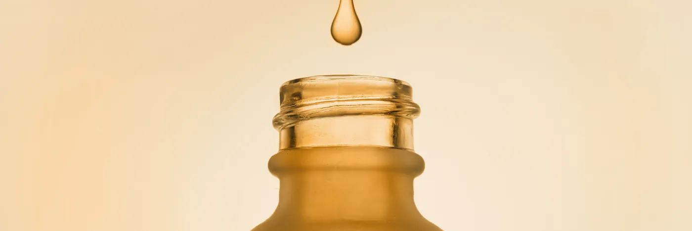 A single golden droplet falling towards the open neck of an unlabelled amber glass bottle against a warm yellow background.