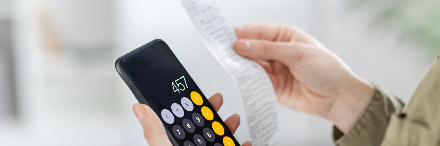 A person holding a calculator in one hand and a long paper receipt in the other, working through an itemised list of expenses.