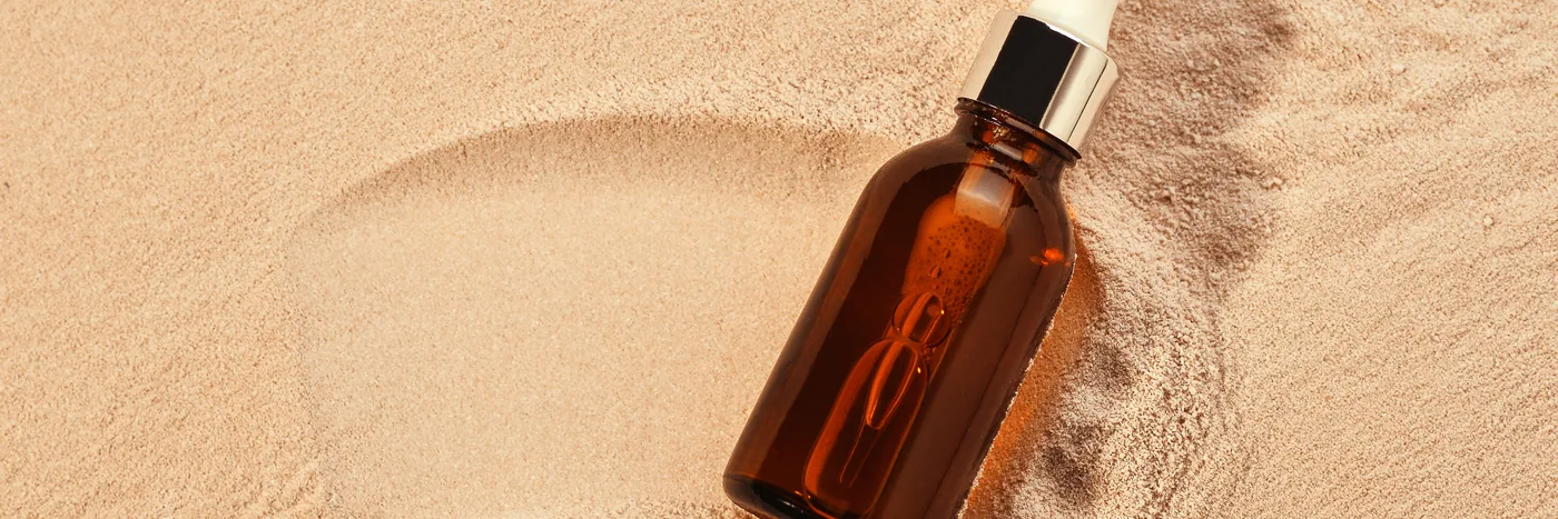 An unlabelled amber glass serum bottle resting in a smooth swirl of pale sand.